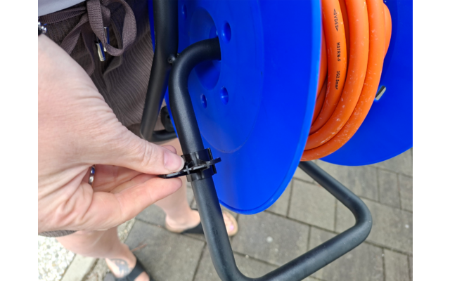 Hand hält einen schwarzen Kabelbinder um das schwarze Metallgestell einer blauen Kabeltrommel mit orangefarbenem Kabel; Pflasterboden und Beine mit Sandalen und Shorts sichtbar; Rucksackriemen am Rand