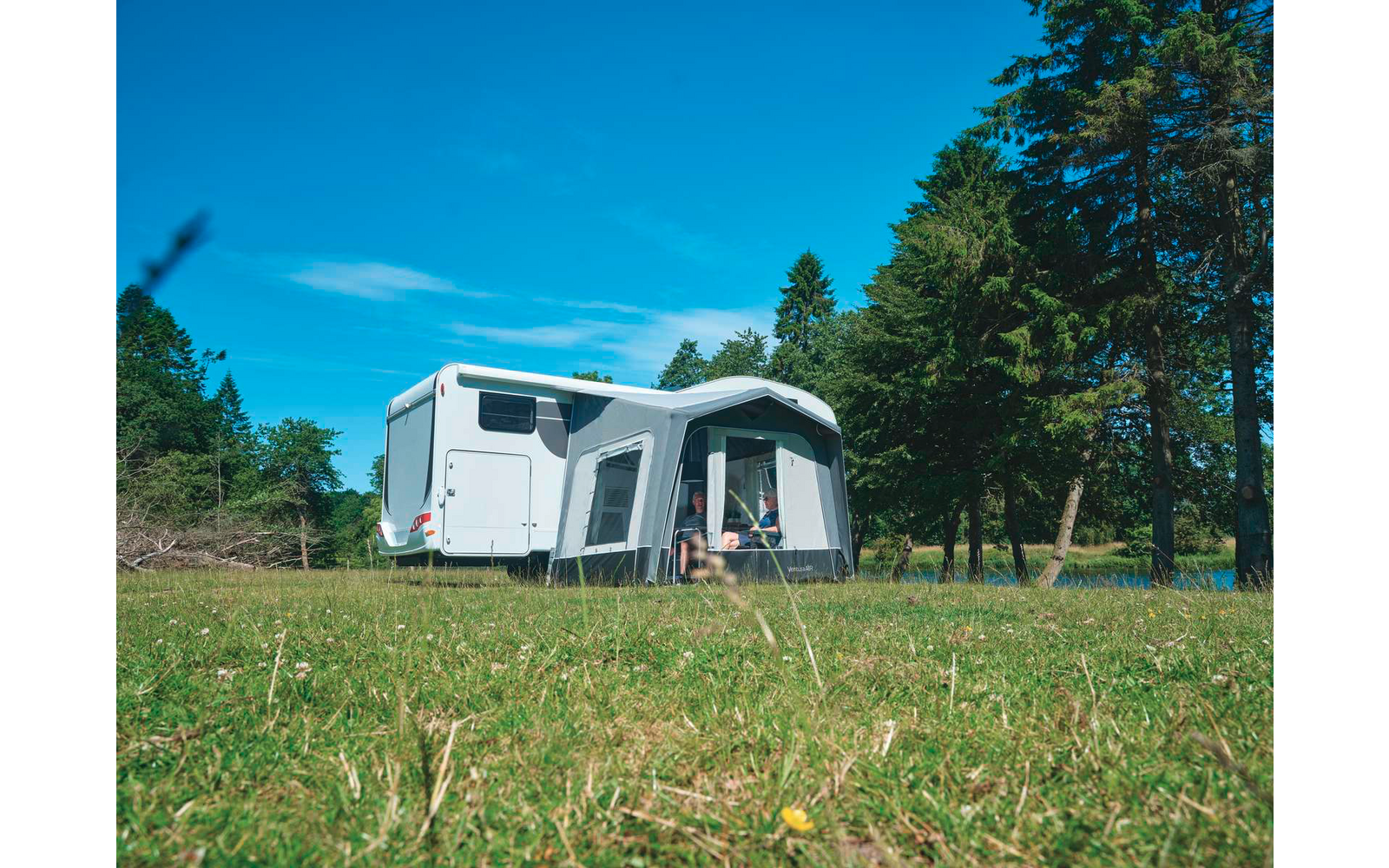 Weißes Wohnmobil mit angebautem Vorzelt auf einer Wiese, Bäume im Hintergrund und blauer Himmel