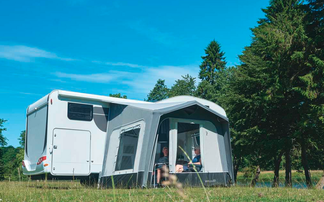 Autocaravana blanca con toldo anexo en un campo de hierba, árboles al fondo y cielo azul