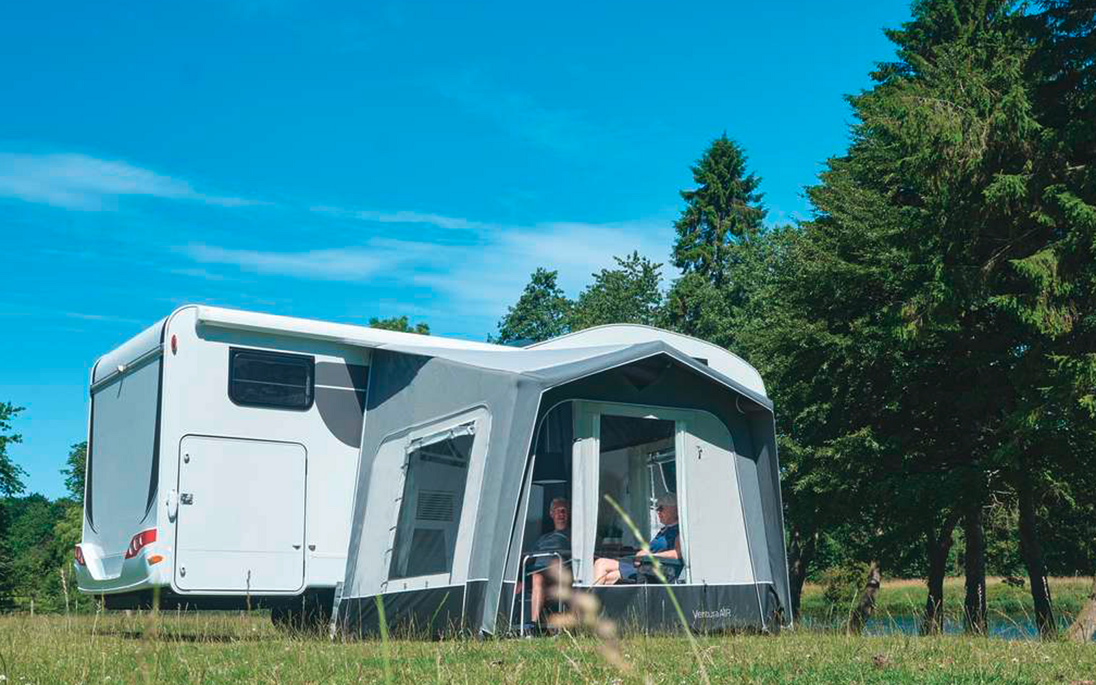 Autocaravana blanca con toldo anexo en un campo de hierba, árboles al fondo y cielo azul