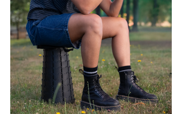 Person sitzt auf einem schwarzen Hocker im Freien, trägt blaue Shorts, schwarze Socken und schwarze Schuhe