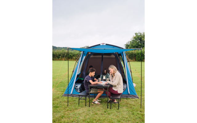 Drei Personen sitzen an einem kleinen Campingtisch vor einem geöffneten blauen Zelt auf einer grünen Wiese; auf dem Tisch stehen Teller und Becher