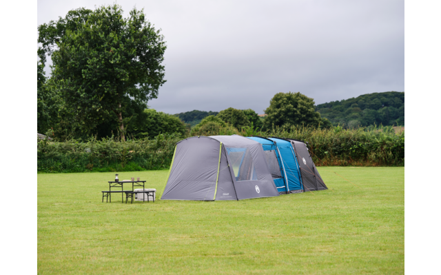 Großes graublaues Zelt auf einer Wiese, links ein Klapptisch und zwei Stühle, im Hintergrund Hecke, Bäume und bewölkter Himmel