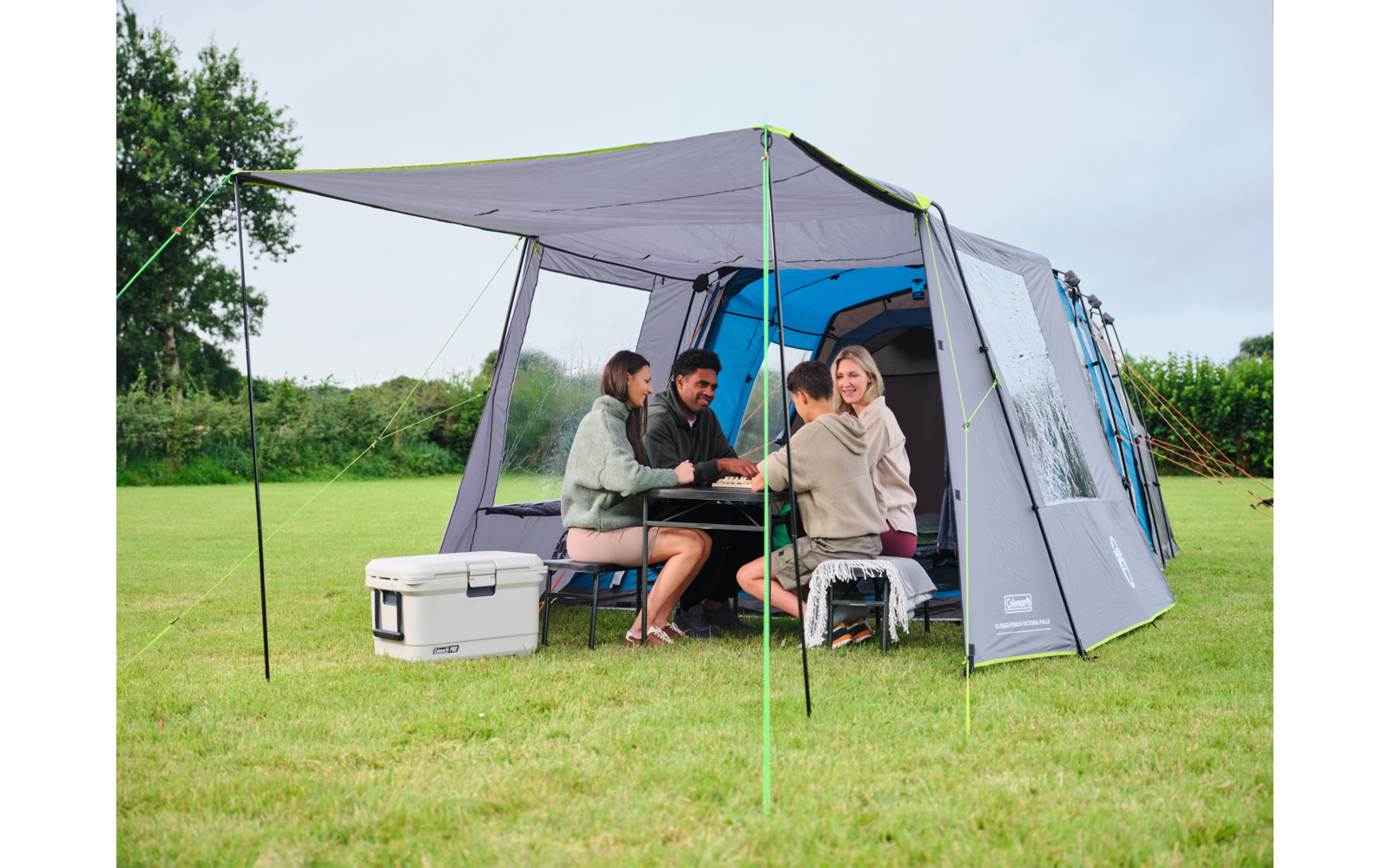 Vier Personen sitzen an einem Klapptisch unter dem Vordach eines grauen Campingzelts auf einer Wiese; Kühlbox und geöffnetes Zelt sichtbar