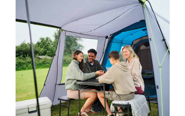 Vier Personen sitzen an einem Klapptisch im Vorzelt eines Campingzelts; im Hintergrund Zeltöffnung und Wiese; Kühlbox links