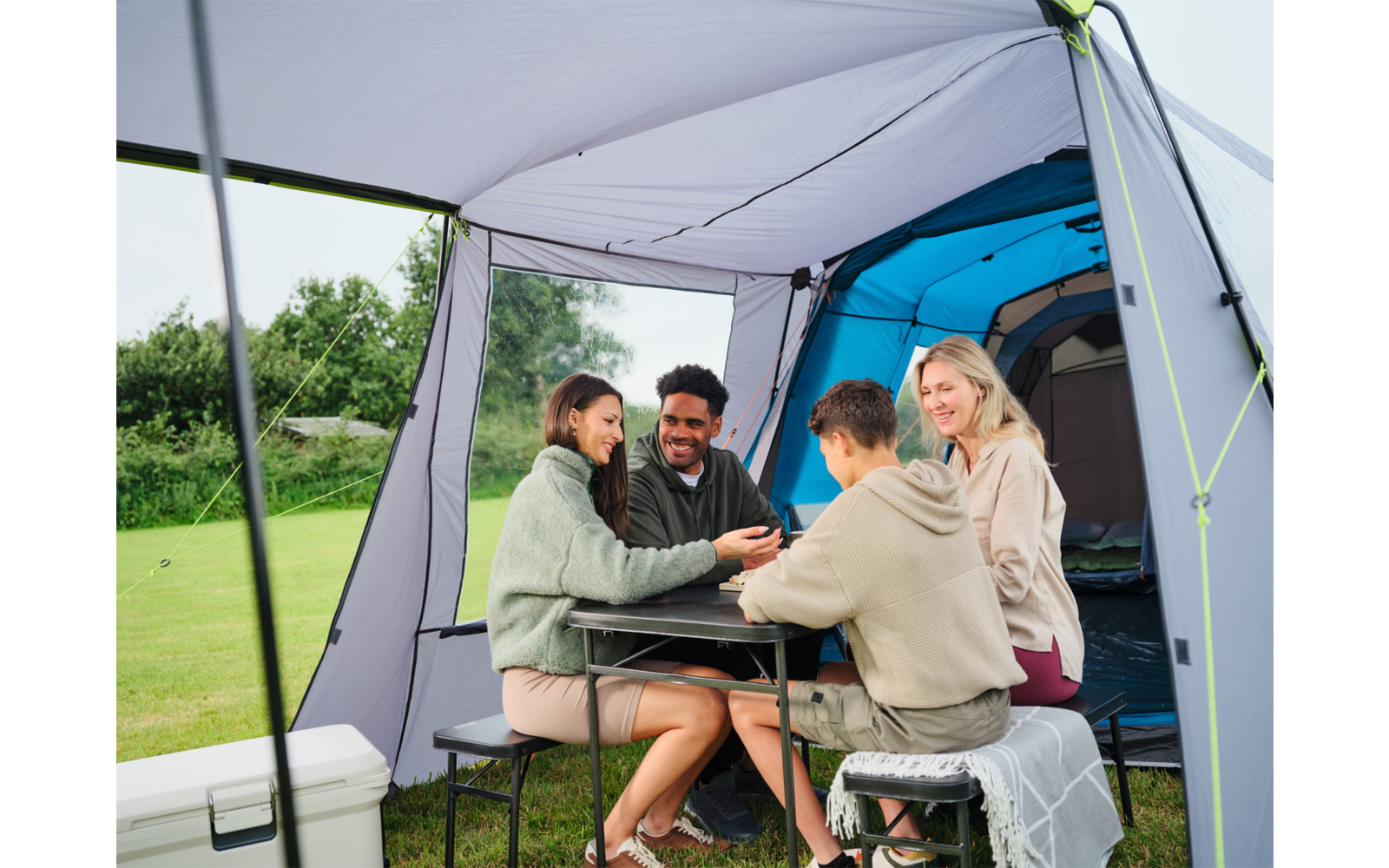 Vier Personen sitzen an einem Klapptisch im Vorzelt eines Campingzelts; im Hintergrund Zeltöffnung und Wiese; Kühlbox links
