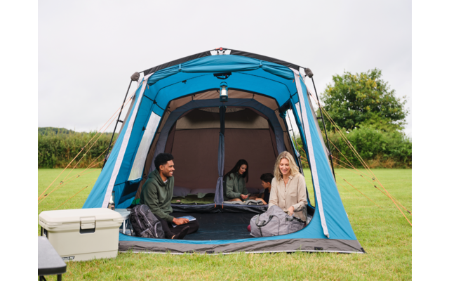 Drei Personen sitzen in einem offenen blauen Zelt auf einer Wiese; links eine weiße Kühlbox; im Hintergrund Bäume und bewölkter Himmel