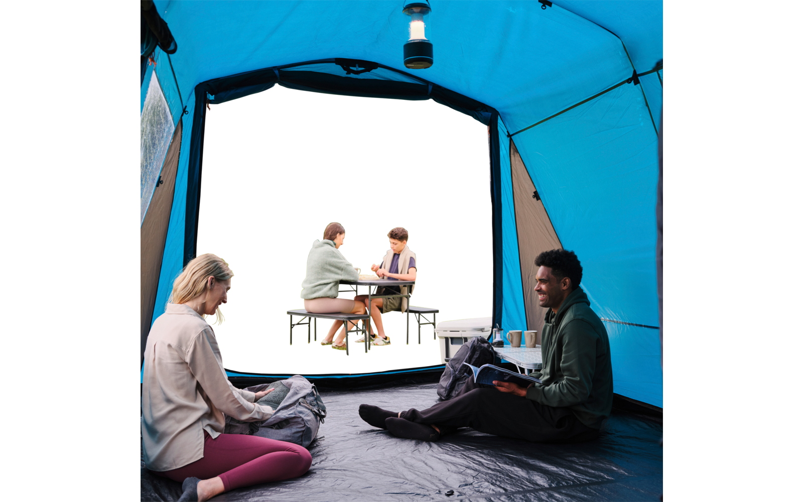 Innenansicht eines blauen Campingzelts: zwei Personen sitzen auf Matten, eine hält ein Buch, die andere etwas in den Händen. An der Zeltdecke hängt eine Laterne. Durch die geöffnete Tür sitzen zwei Personen an einem Picknicktisch; neben dem Eingang s