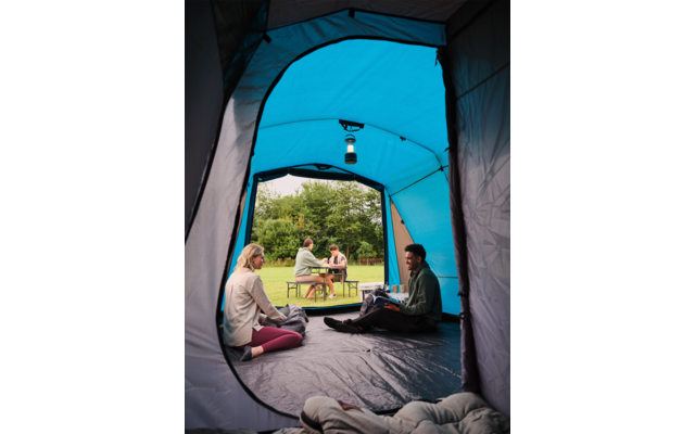 Blaues Zelt mit geöffneter Tür; im Inneren zwei Personen auf dem Boden; Laterne hängt an der Zeltdecke; vor dem Zelt sitzen zwei Personen an einem Picknicktisch auf einer Wiese; Bäume im Hintergrund
