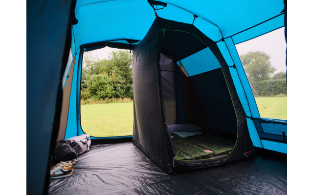 Innenansicht eines blauen Zeltes mit dunklem innerem Schlafabteil, grüner Isomatte und Kissen, Schuhe und Tasche auf dem Boden, geöffnete Tür mit Blick auf Wiese und Bäume