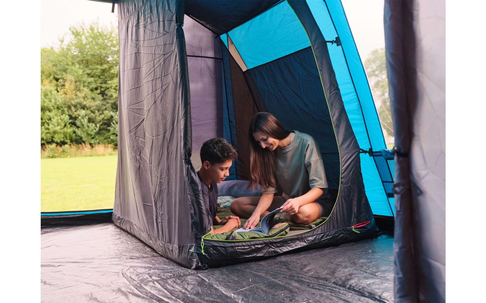Zwei Personen sitzen im Eingang eines blau-grauen Zelts und schauen auf Gegenstände auf einer Isomatte, im Hintergrund Rasen und Bäume