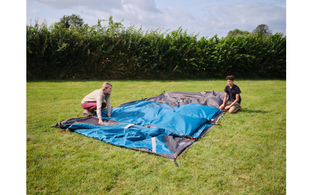 Zwei Personen breiten ein blau-graues Zelt auf einer grünen Wiese aus; Hecke im Hintergrund, Himmel mit Wolken