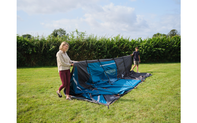 Zwei Personen halten ein teilweise aufgebautes blaues Zelt auf einer Wiese, im Hintergrund eine Hecke und ein leicht bewölkter Himmel