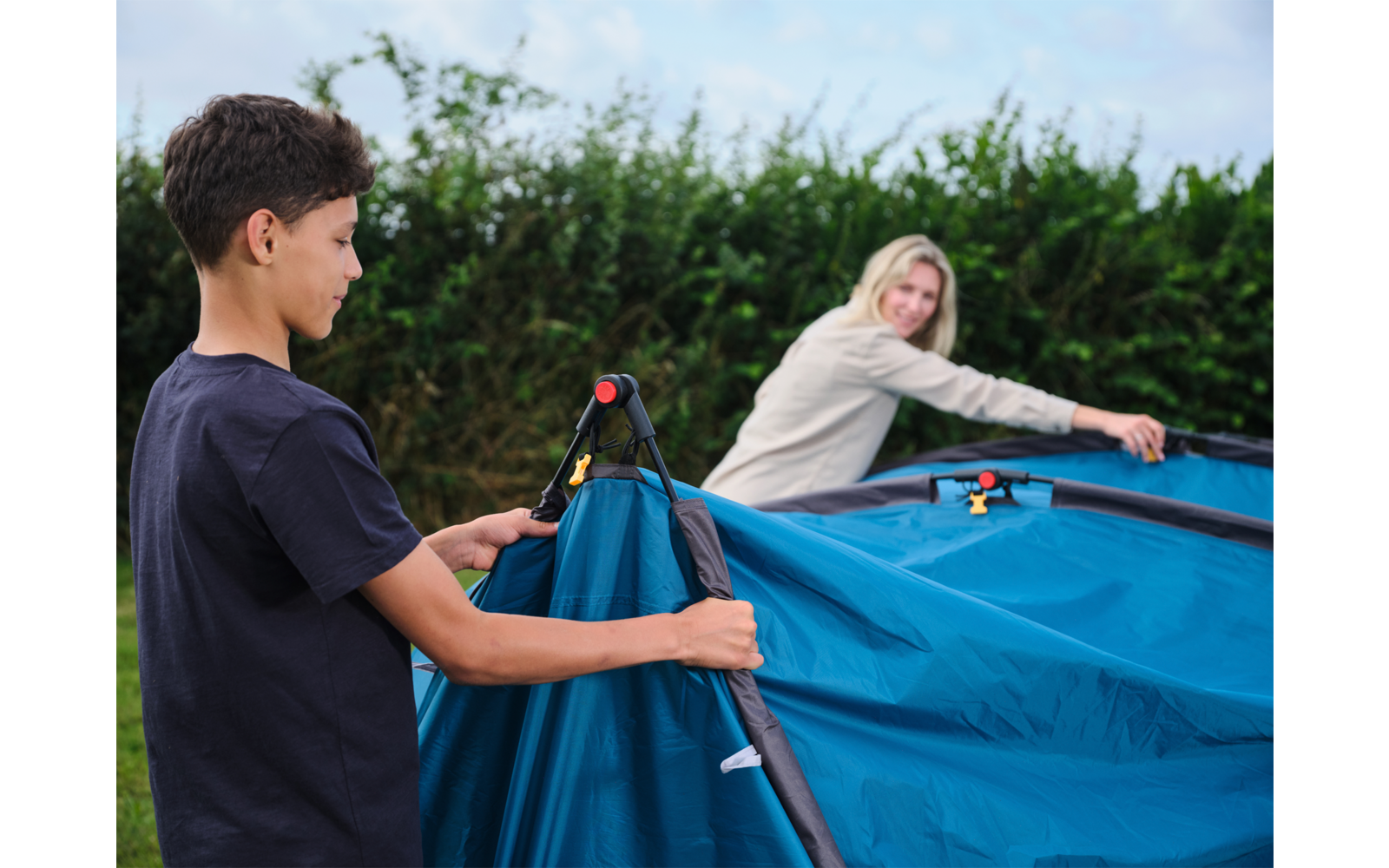 Zwei Personen, ein Junge links und eine Frau rechts, halten Zeltstangen und spannen ein blaues Zelt vor einer Hecke