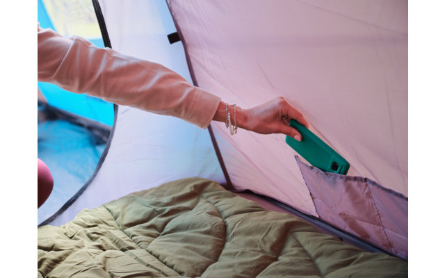 Hand mit Armbändern hält ein grünes Smartphone über eine Innentasche an der Zeltwand; grüner Schlafsack liegt im Zelt