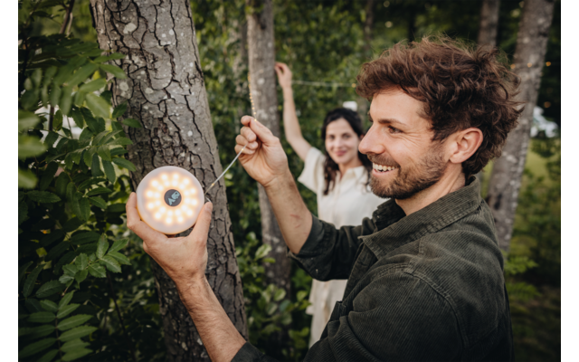 Man hangt een ronde lamp aan een boom, vrouw staat achterin en houdt een snoer vast, bomen en groen op de achtergrond