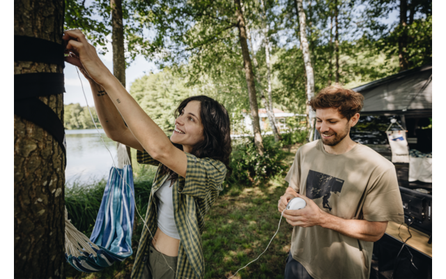 Vrouw bevestigt gestreepte hangmat aan een boom, man houdt touw vast, bomen en meer op de achtergrond