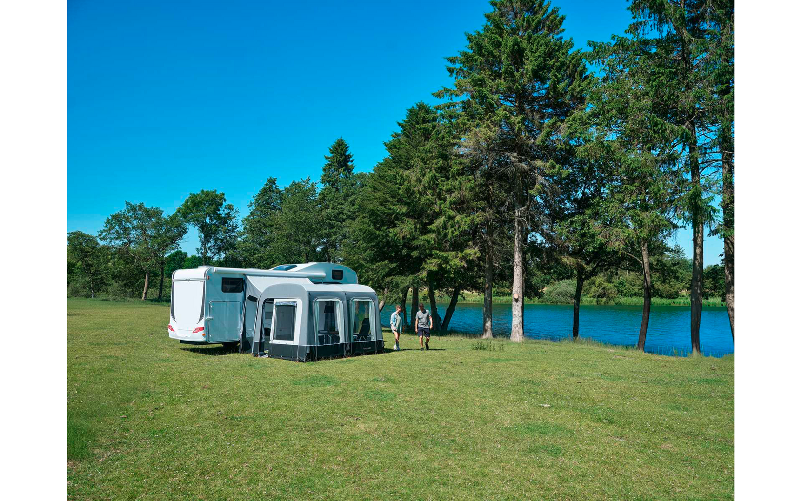 Weißer Wohnwagen mit grauem Vorzelt auf einer Grasfläche neben Bäumen und einem See, zwei Personen gehen am Ufer, blauer Himmel