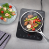 Inductiekookplaat met koekenpan met garnalen, gebruinde vleesplakjes, groene asperges en gehalveerde cherrytomaten; daarnaast een bord met garnalen en asperges en een geruit keukendoek