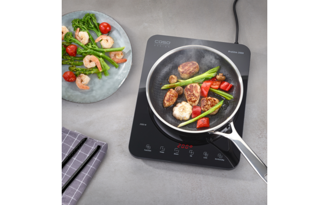 Inductiekookplaat met koekenpan met garnalen, gebruinde vleesplakjes, groene asperges en gehalveerde cherrytomaten; daarnaast een bord met garnalen en asperges en een geruit keukendoek