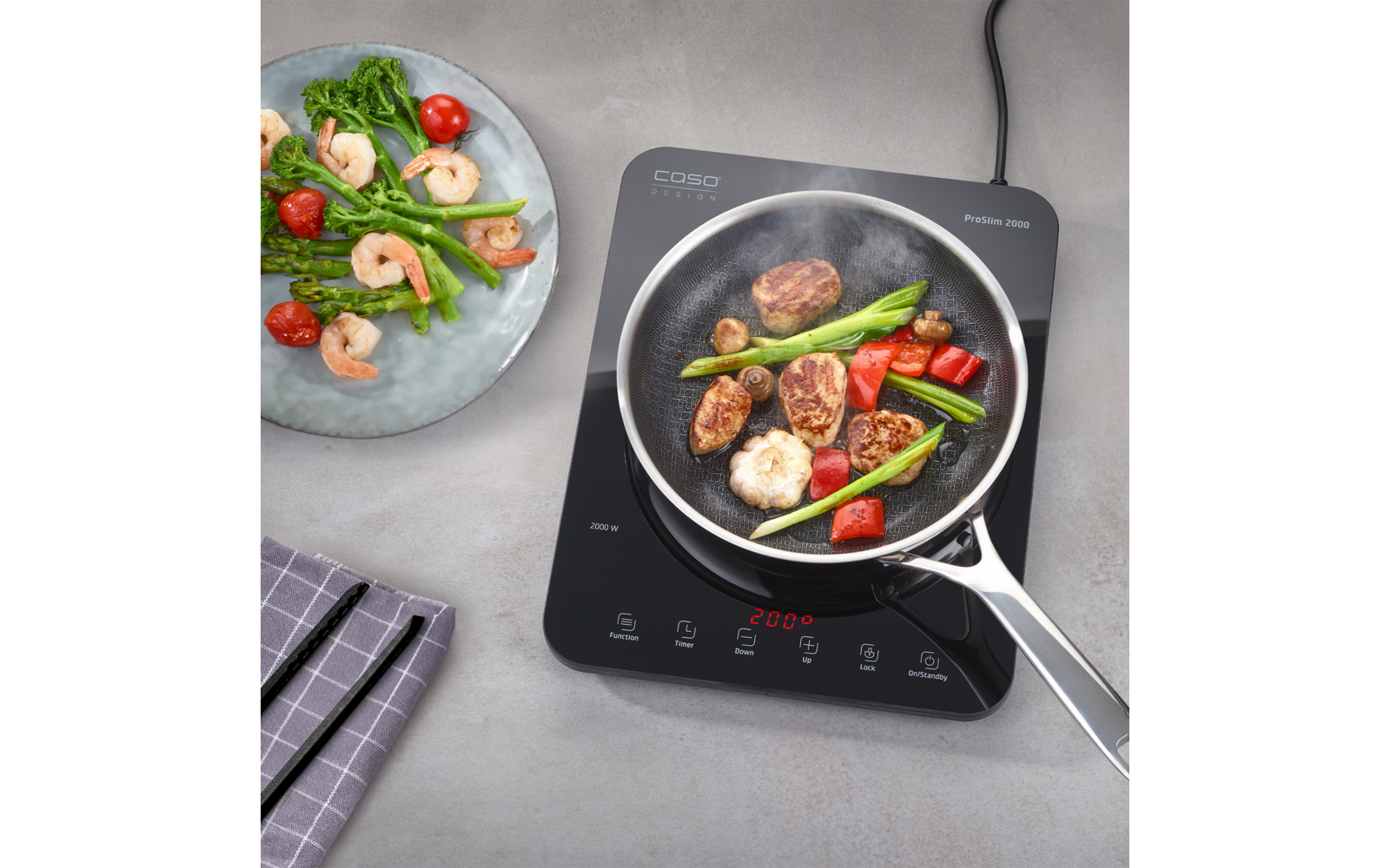 Inductiekookplaat met koekenpan met garnalen, gebruinde vleesplakjes, groene asperges en gehalveerde cherrytomaten; daarnaast een bord met garnalen en asperges en een geruit keukendoek