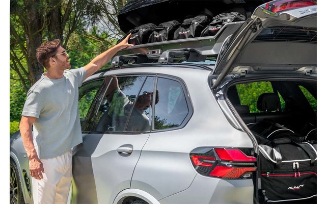 Silbernes SUV mit geöffneter Heckklappe und geöffneter Dachbox; eine Person steht seitlich und greift in die Dachbox; mehrere Helme in der Dachbox sichtbar; Reisetaschen im Kofferraum; Bäume im Hintergrund
