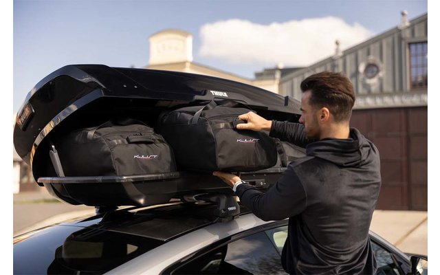 Person legt schwarze Reisetaschen in einen geöffneten Dachkoffer auf einem Auto; Gebäude und Himmel im Hintergrund