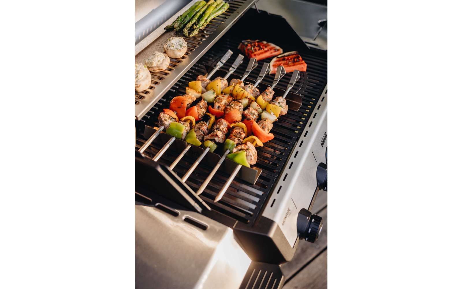 Gasgrill mit Metallrost, mehreren Spießen mit Fleischstücken, Paprika, Zwiebeln und Kartoffelwürfeln; Pilze, grüner Spargel und halbierte Tomaten auf dem Grill