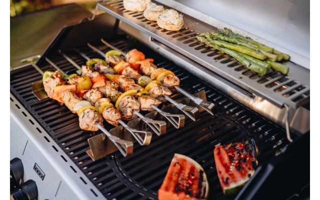 Gasgrill mit mehreren Metallspießen mit Fleisch- und Gemüsestücken; oben rechts gebündelte grüne Spargelstangen; oben links belegte runde Brotscheiben; unten rechts gegrillte Melonenstücke