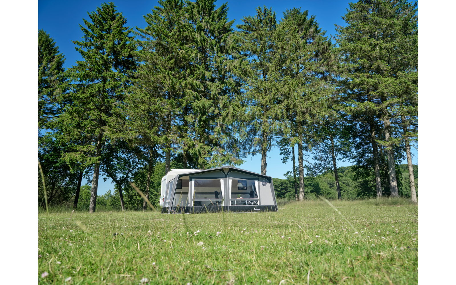 Weißer Wohnwagen mit angebautem Vorzelt auf grüner Wiese, im Hintergrund hohe Nadelbäume und blauer Himmel
