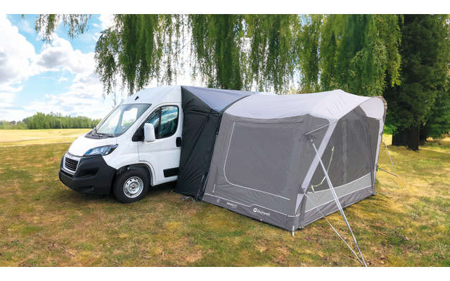 Witte camper met grijs opblaasbare voortent op een grasveld, bomen en bewolkte lucht op de achtergrond