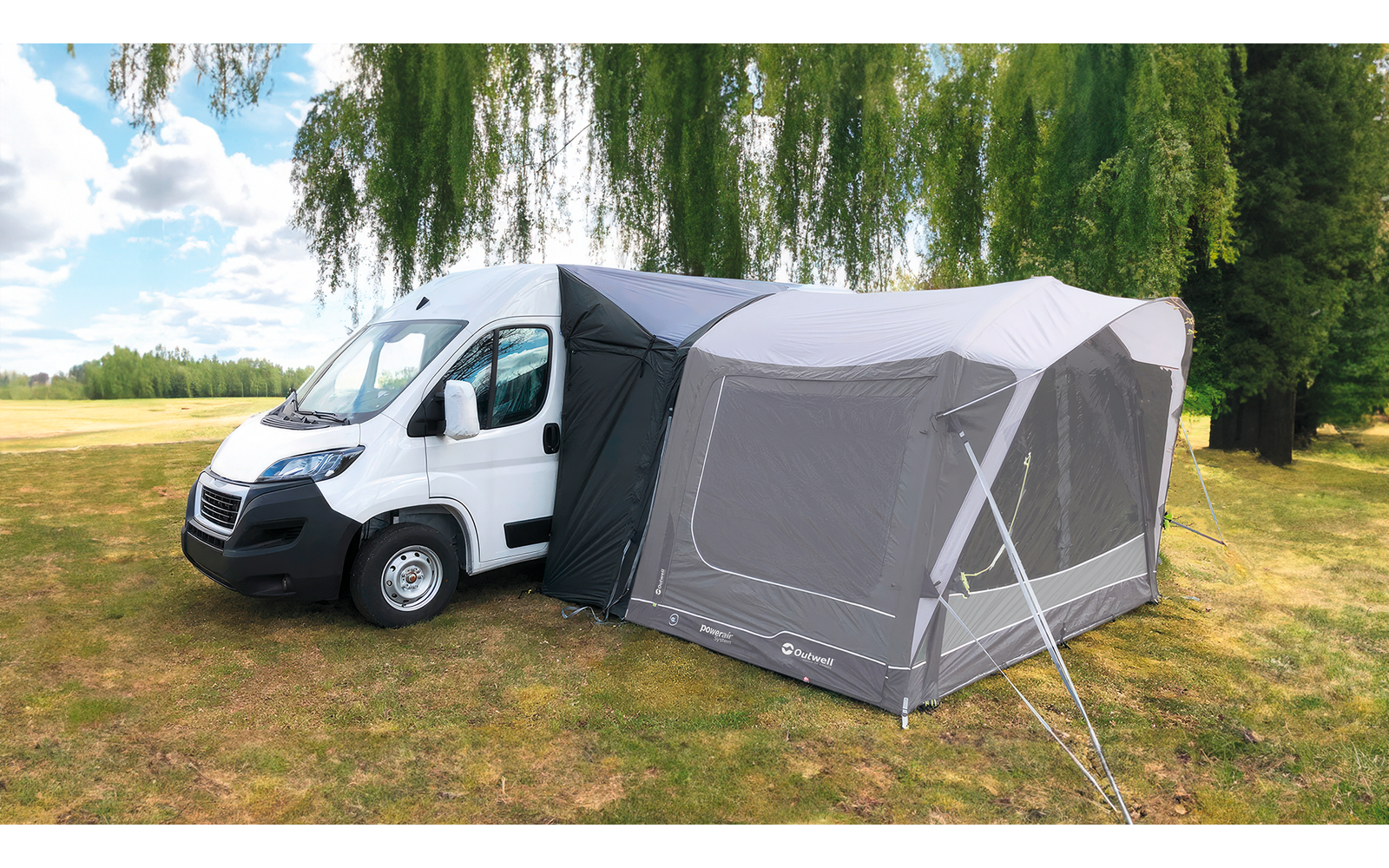 Witte camper met grijs opblaasbare voortent op een grasveld, bomen en bewolkte lucht op de achtergrond