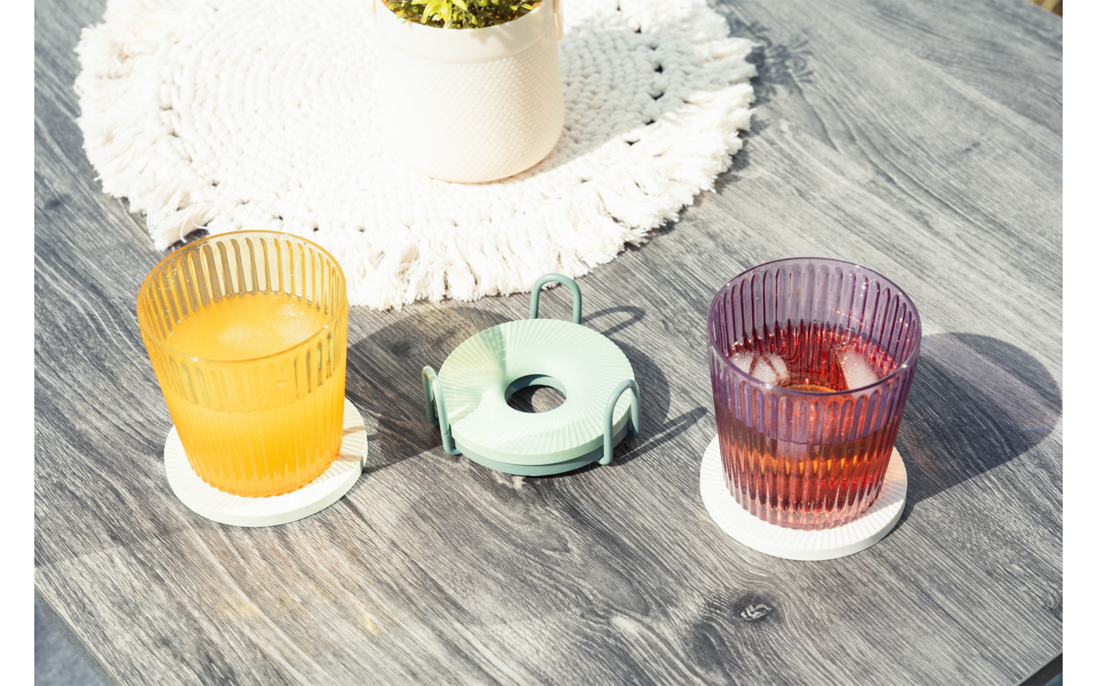 Twee geribbelde glazen op witte onderzetters op een grijze houten tafel, links glas met oranje vloeistof, rechts glas met roodbruine vloeistof en ijsblokjes; daartussen een rond groen voorwerp met een centrale opening en metalen draad; bovenaan een wit gehaakt onderzetter met een klein plantenpotje