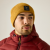 Hombre con gorro de punto amarillo mostaza con logo cuadrado negro y cazadora acolchada granate con capucha, mira hacia la derecha, fondo claro