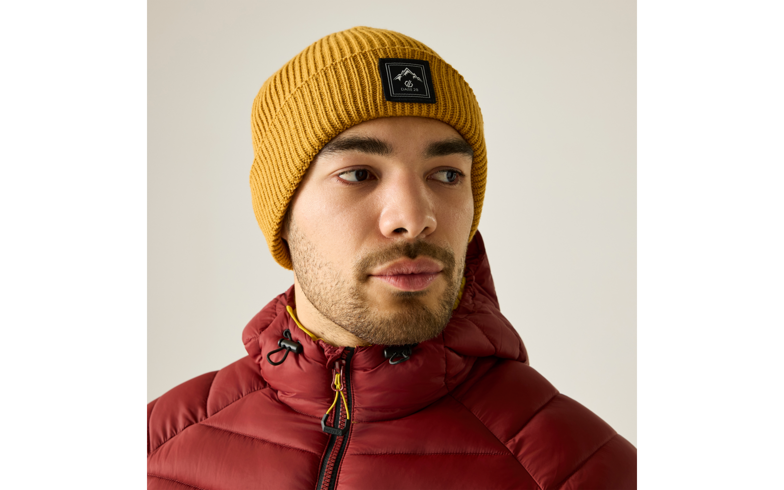 Hombre con gorro de punto amarillo mostaza con logo cuadrado negro y cazadora acolchada granate con capucha, mira hacia la derecha, fondo claro