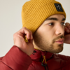 Primer plano de una persona que lleva un gorro de punto amarillo con logo negro y una chaqueta acolchada roja, ajustándose el gorro con ambas manos, mirando hacia la derecha sobre fondo beige