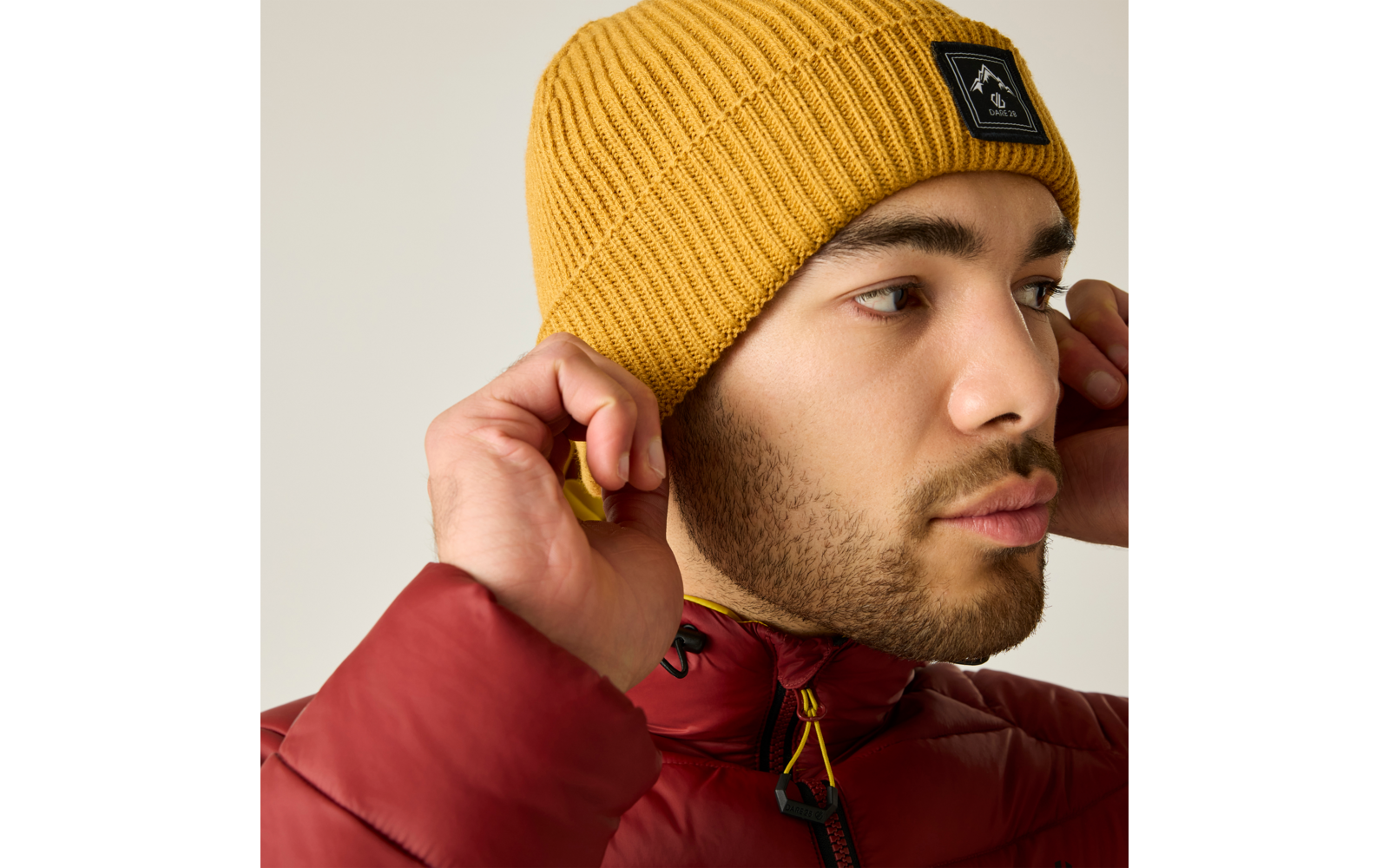 Primer plano de una persona que lleva un gorro de punto amarillo con logo negro y una chaqueta acolchada roja, ajustándose el gorro con ambas manos, mirando hacia la derecha sobre fondo beige