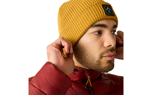 Gros plan d'une personne portant un bonnet en maille jaune avec un logo noir et une doudoune rouge, ajustant le bonnet des deux mains, regard vers la droite sur fond beige