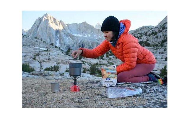 Person in orange Daunenjacke und schwarzer Mütze kniet auf Fels und hält einen Topf über einem kleinen Gaskocher; Metalltasse, Trockenfutterbeutel und Berggipfel im Hintergrund