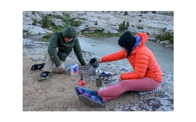 Two people sit on a rocky lakeshore; one pours liquid from a cup into a pot on a small gas stove; backpacks and gear lie beside them