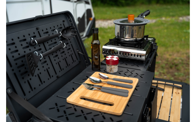 Gasfornuis met afgedekte pan, fles olie, pot met rood-wit geruit deksel, snijplank met vork, lepel en mes op een zwart metalen oppervlak; gedeeltelijk zicht op een witte bestelwagen op de achtergrond