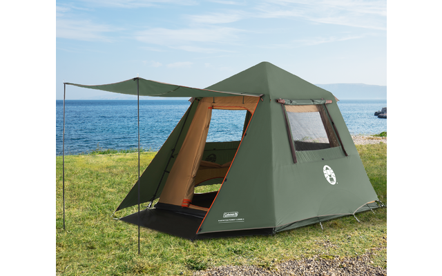 Green camping tent with open entrance and awning on grass; sea in the background, rocks to the right, blue sky with clouds