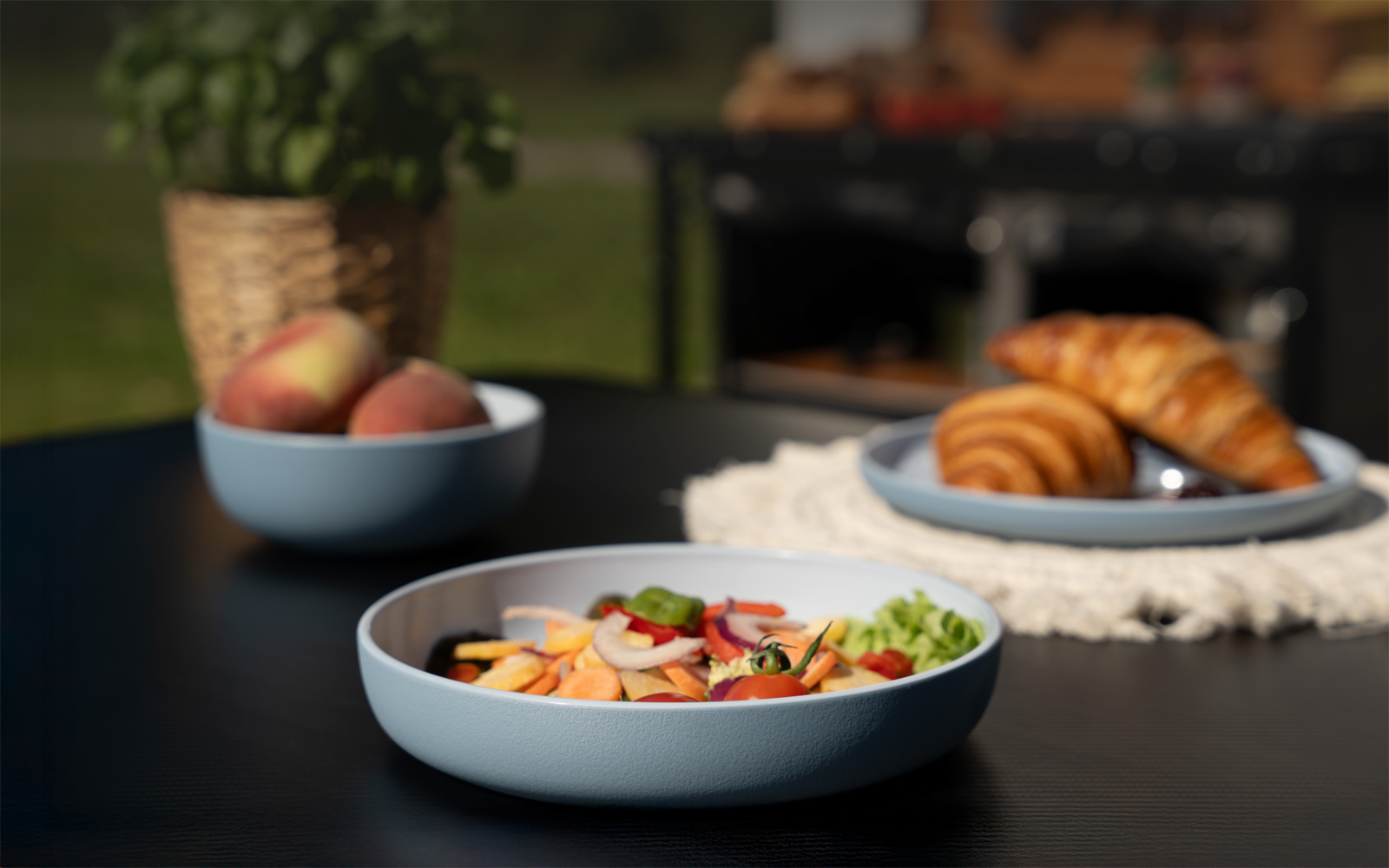 Blaue Schale mit geschnittenem Obst auf einem Tisch; dahinter Teller mit Croissants, Schüssel mit ganzen Pfirsichen und ein Topf mit Pflanze; unscharfer Grill im Hintergrund