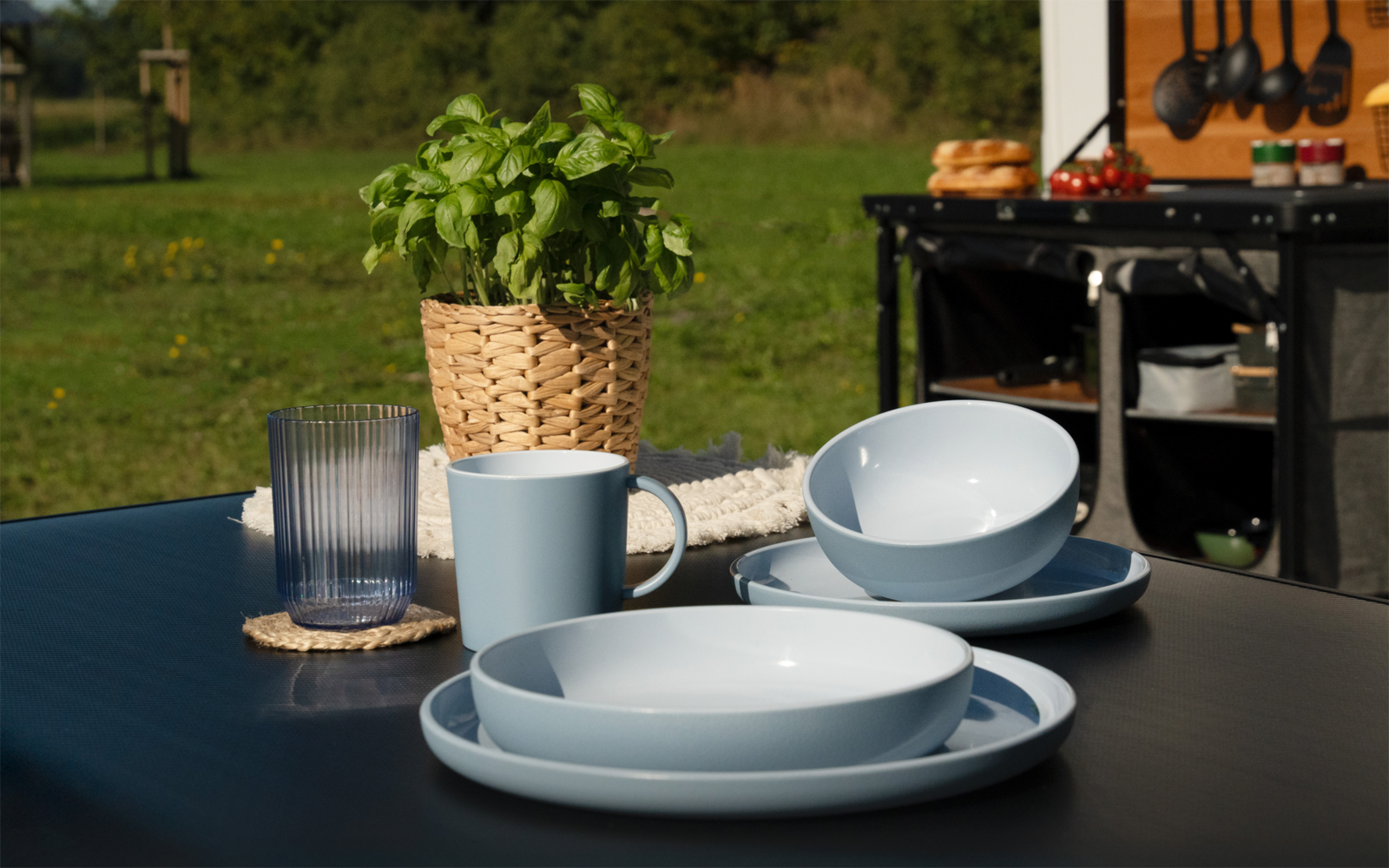 Schwarzer Gartentisch mit hellblauen Tellern, Schüssel und Tasse, Glas auf Untersetzer, Basilikum im geflochtenen Korb; im Hintergrund Arbeitsfläche mit aufgehängten Kochutensilien und Rasen