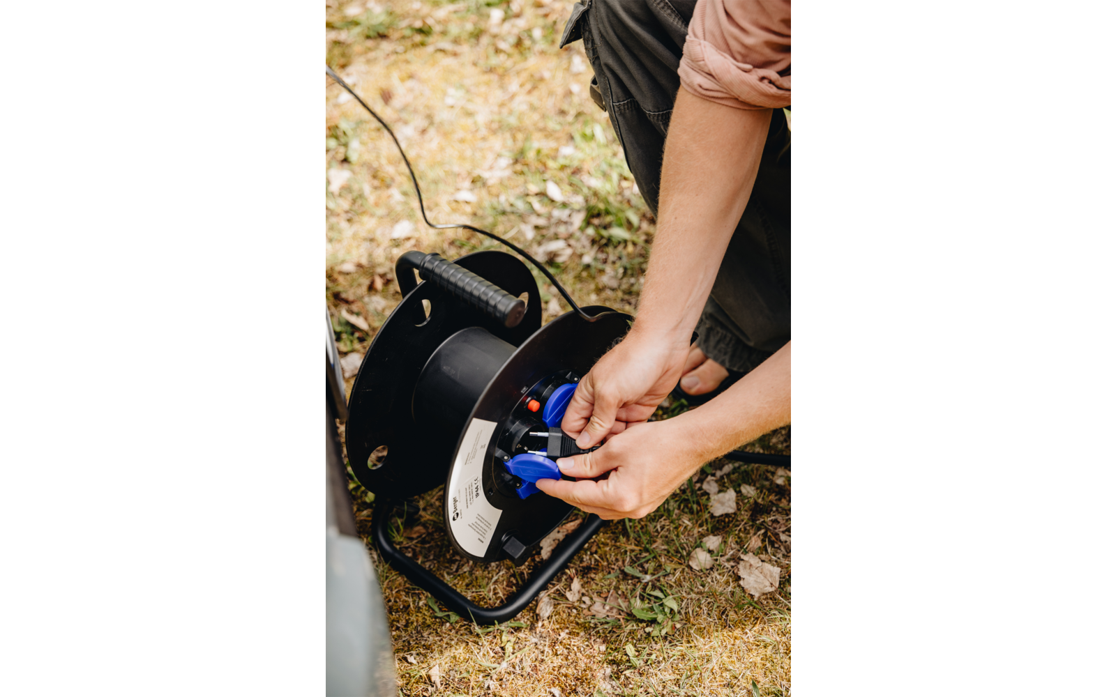 Des mains insèrent un connecteur bleu dans une bobine de câble noire sur de l'herbe sèche; pantalon et manche retroussée visibles