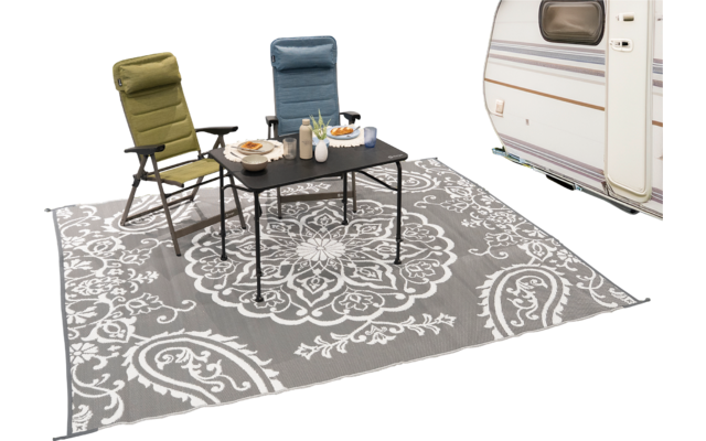 Zwei Campingstühle (grün, blau) und ein schwarzer Klapptisch mit Tellern, Flaschen und Schalen auf einem grauen Teppich mit weißem Ornamentmuster; rechts teilweise ein Wohnwagen sichtbar