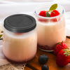 Deux petits pots en verre avec une crème blanche et une couche de fruits rouges en bas ; un pot avec un couvercle noir, l'autre ouvert garni de framboises et d'une petite feuille verte ; fraises et myrtilles à côté des pots sur une planche en bois ; tissu blanc en arrière-plan