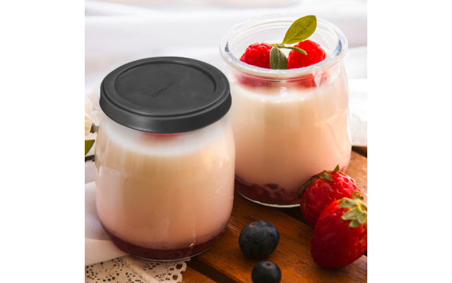 Deux petits pots en verre avec une crème blanche et une couche de fruits rouges en bas ; un pot avec un couvercle noir, l'autre ouvert garni de framboises et d'une petite feuille verte ; fraises et myrtilles à côté des pots sur une planche en bois ; tissu blanc en arrière-plan