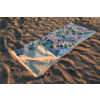 Une serviette de plage colorée à motif déroulée sur du sable ; un coin replié montre l'envers blanc en éponge ; sable avec sillons et empreintes de pas