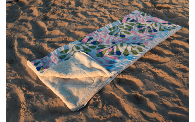 Une serviette de plage colorée à motif déroulée sur du sable ; un coin replié montre l'envers blanc en éponge ; sable avec sillons et empreintes de pas